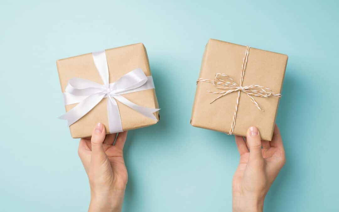 First person top view photo of female hands holding two craft paper gift boxes with twine bow and white satin ribbon bow on isolated pastel blue background